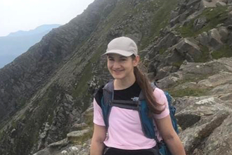 Daisy Askari sitting in mountains, wearing baseball cap, pink T-shirt, dark trousers and climbing boots