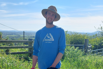 Joe Franklin with his dog on a summer's day, wearing blue top and beige hat.  Background of green grass and blue sky.