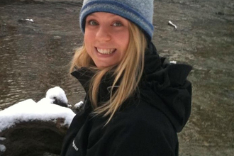 Lucy Boyett, near water in winter, wearing grey woollen beanie hat and black jacket with hood 