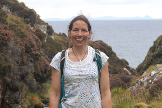 Rhian Hughes outdoors at coast, smiling and wearing a pale top dark trousers and rucksack.