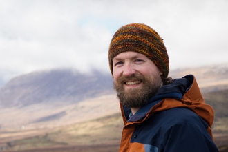 Tim Yardley in mountain setting, wearing an orange beanie hat and orange and blue waterproof jacket. 