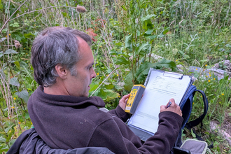 Richard Cutts carrying out ecological survey work, sitting down, marking on a form, holding a bat detector.