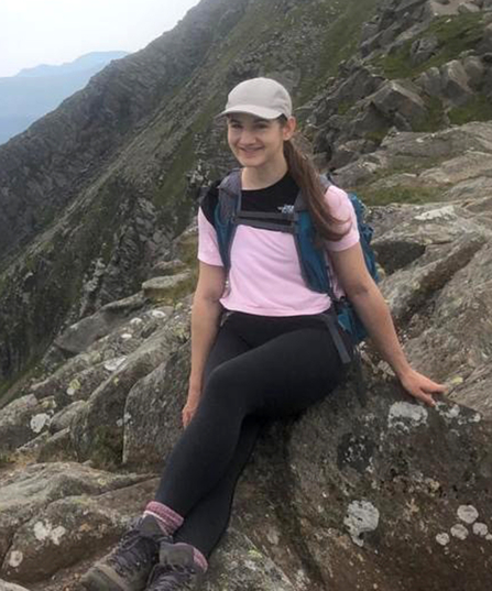 Daisy Askari sitting in mountains, wearing baseball cap, pink T-shirt, dark trousers and climbing boots