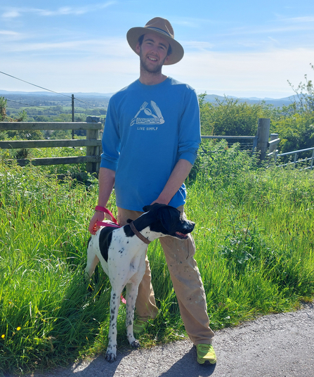 Joe Franklin with his dog on a summer's day, wearing blue top and beige hat.  Background of green grass and blue sky.