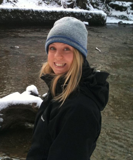 Lucy Boyett, near water in winter, wearing grey woollen beanie hat and black jacket with hood 
