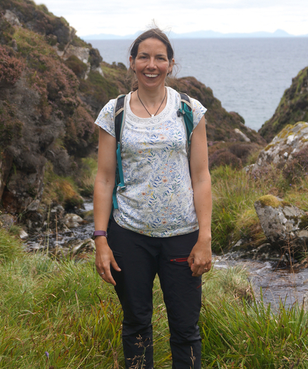 Rhian Hughes outdoors at coast, smiling and wearing a pale top dark trousers and rucksack.