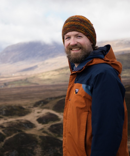 Tim Yardley in mountain setting, wearing an orange beanie hat and orange and blue waterproof jacket. 