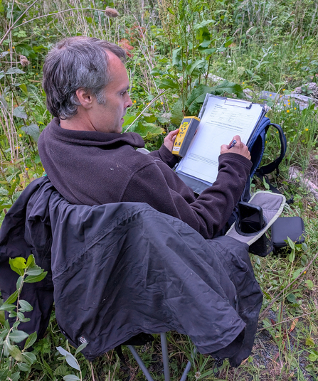 Richard Cutts carrying out ecological survey work, sitting down, marking on a form, holding a bat detector.