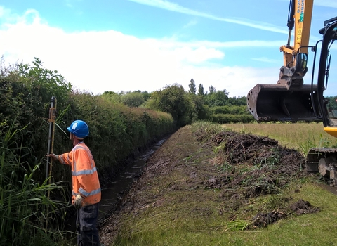 Enfys staff member wearing bright orange hazard jacket, in ditch with digger machinery 